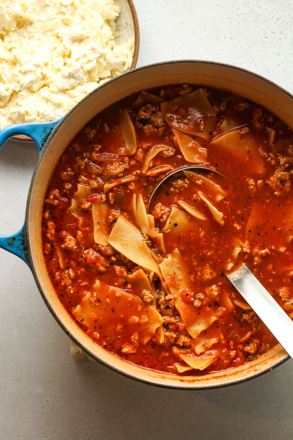 A large pot of turkey lasagna soup with a bowl of 3-cheese blend.