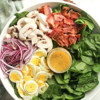 A bowl of spinach salad with eggs, bacon, mushrooms and red onion.