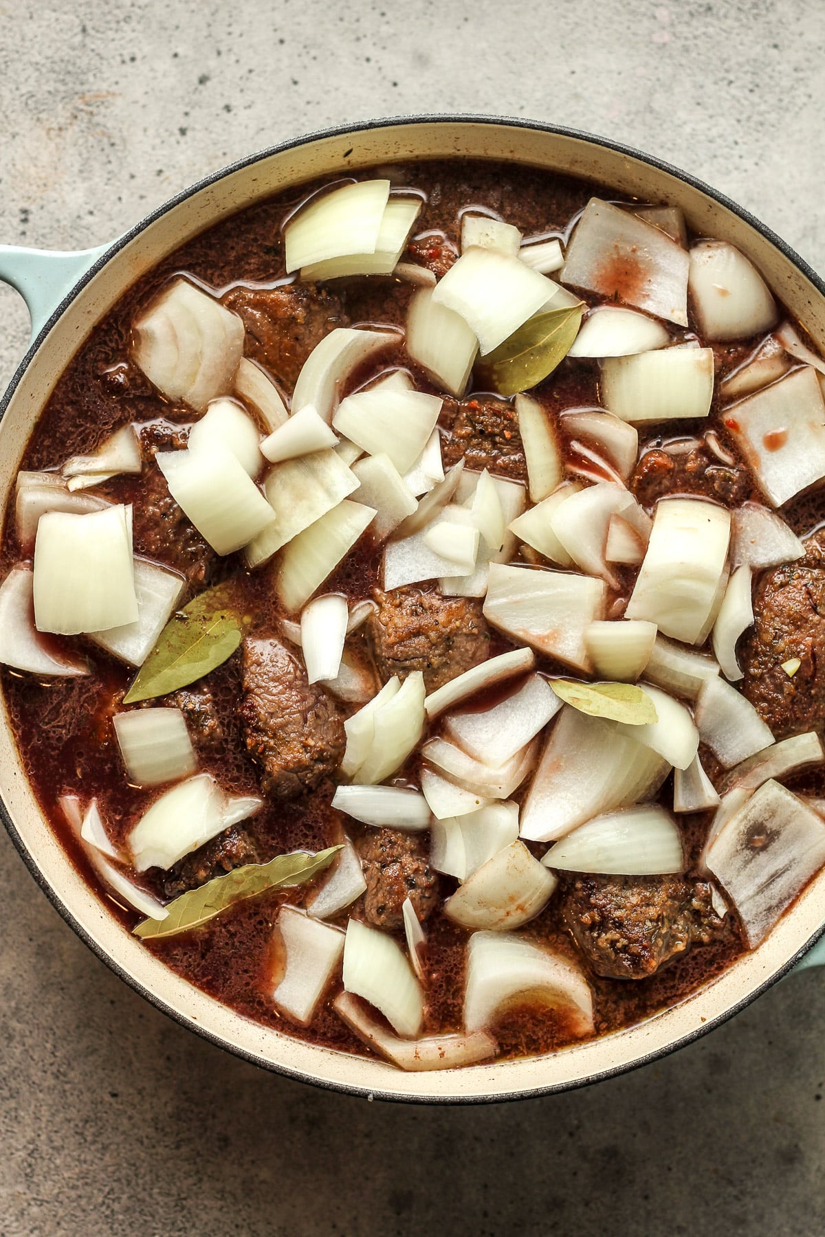 A braiser with the seared beef chunks plus broth, red wine, chunks of onion, and bay leaves.