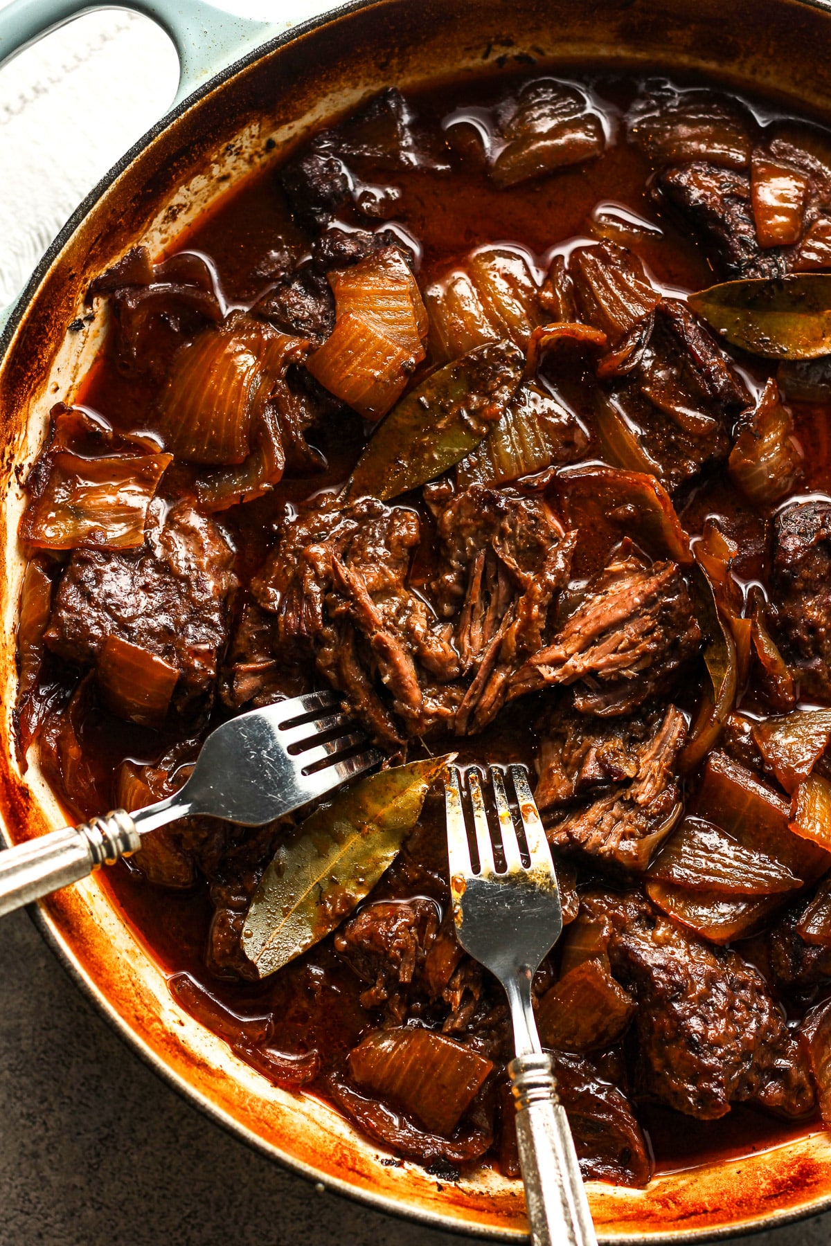 Closeup on a braiser with braised pot roast in a red wine gravy and two forks shredding the beef.