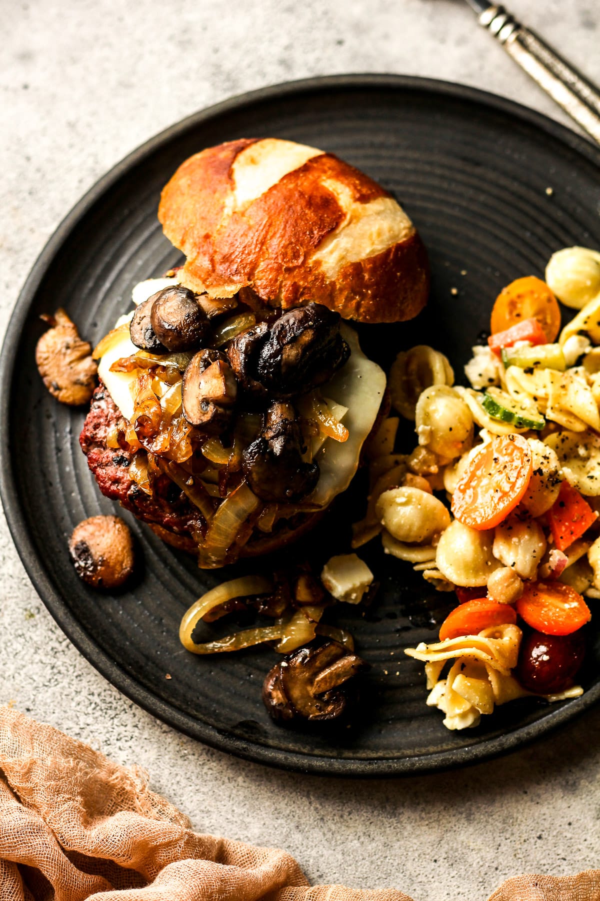 Overhead view of a mushroom swiss burger on a black plate with pasta salad.