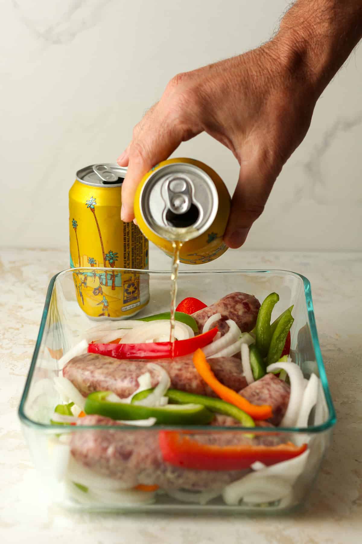 A hand pouring the beer into the dish of brats, onions, and bell peppers.