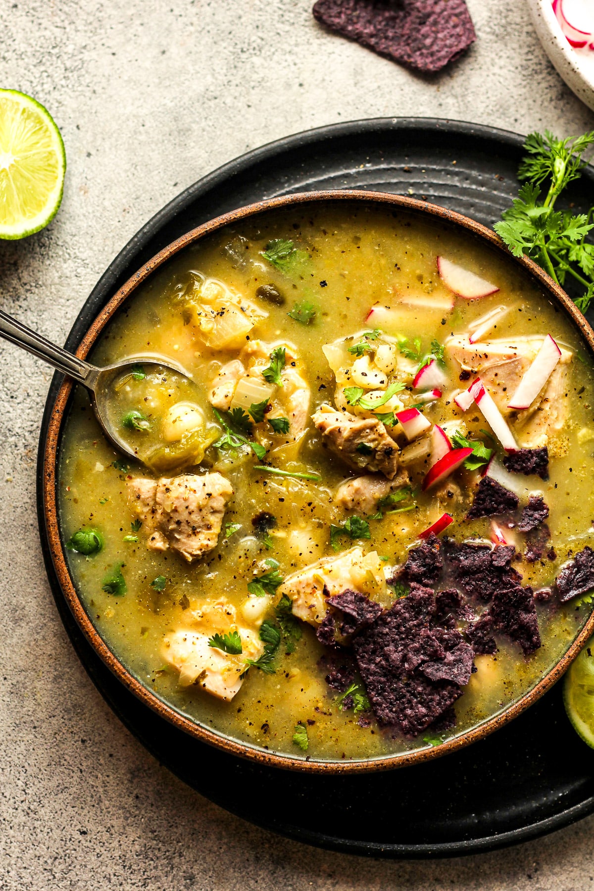 A black bowl of pozole verde with chicken plus a spoon.