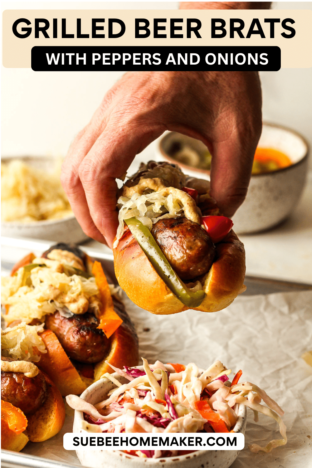 Side view of a hand holding a grilled beer brat with peppers and onions over a tray of brats.