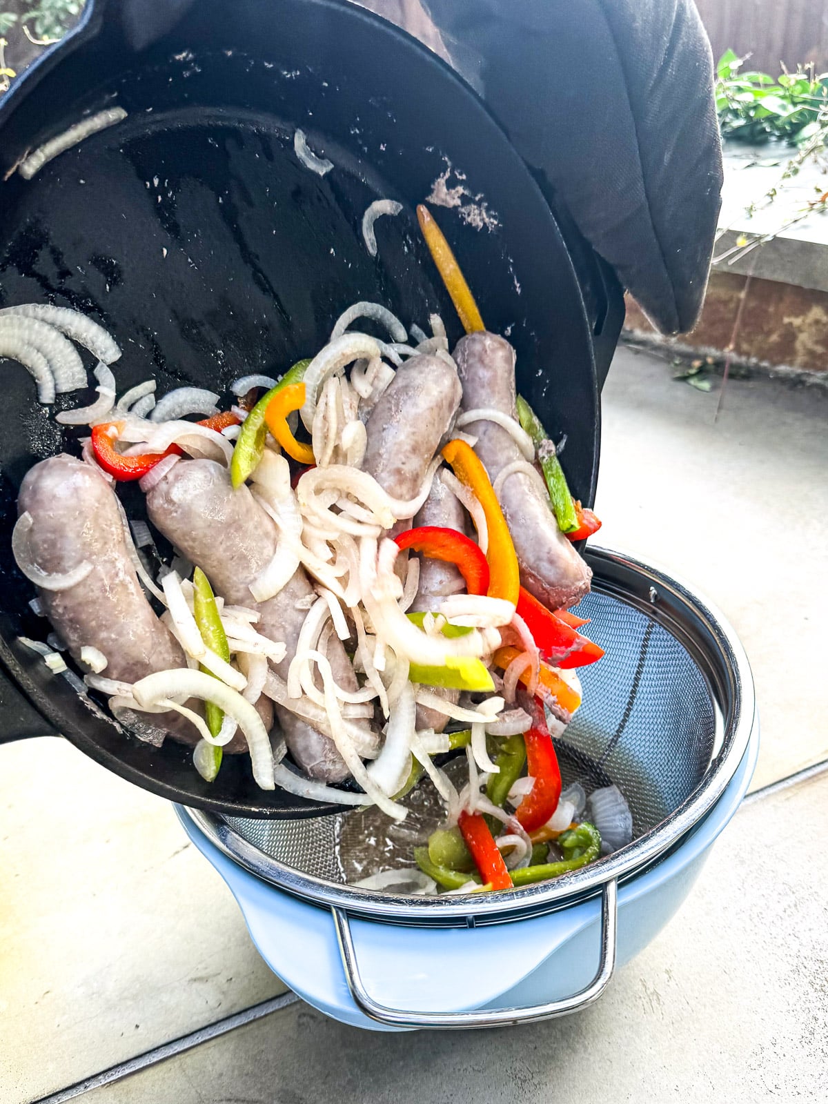 Gloves pouring the boiled brats and veggies into a colander over a bowl.