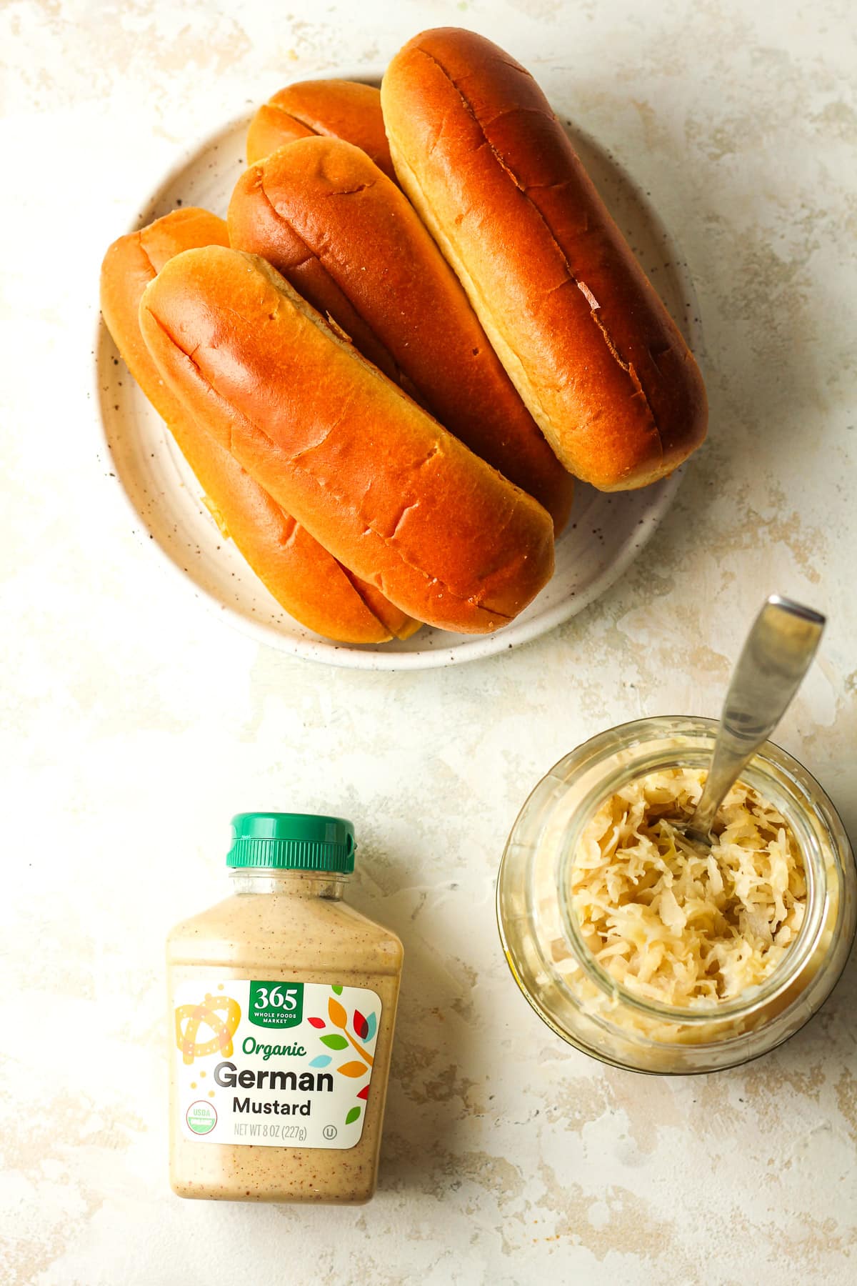 A plate of brioche hot dog buns, a jar of sauerkraut, and a bottle of spicy mustard.