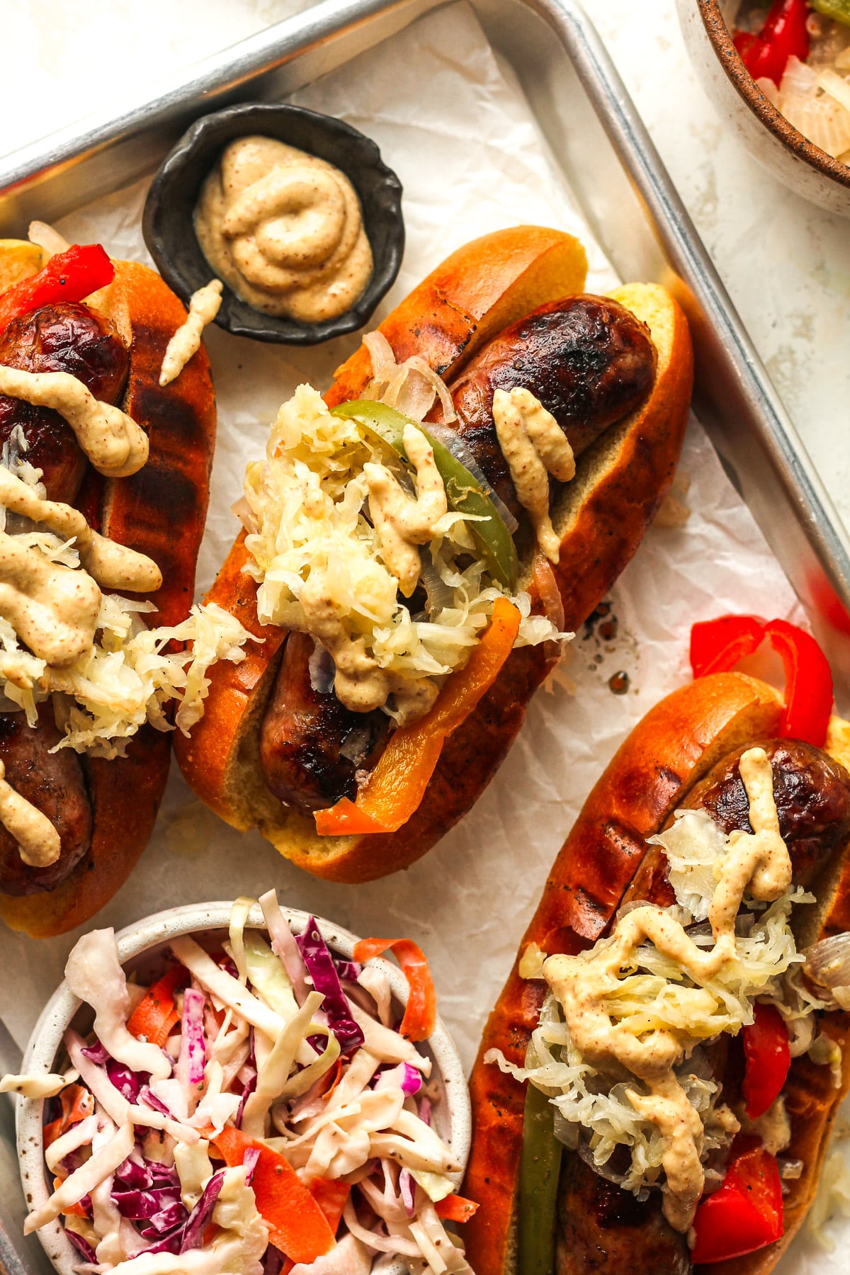 Overhead view of a pan with three grilled beer brats in hotdog buns, with toppings.
