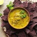 A bowl of salsa verde with some black chips.