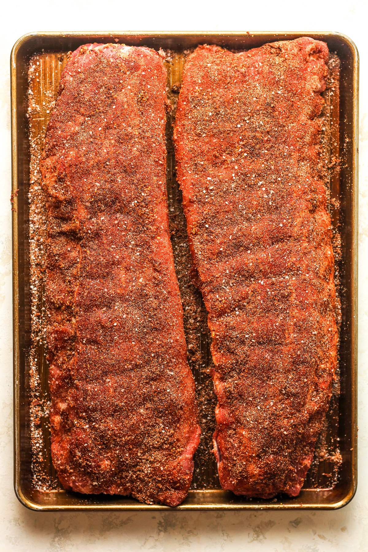 Overhead view of a sheet pan with two racks of the ribs after adding seasoning.